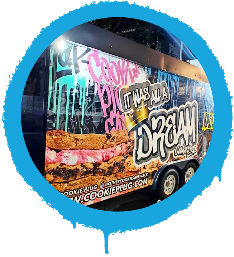 Food truck with colorful graffiti along the side that says 'Cookie Plug' and 'It was all a dream' with a golden crown on top with large cookies in the background.