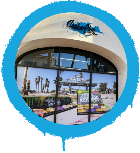 White storefront with a graffiti paint blue splash with a black sign on top that says 'Cookie Plug' with pictures of large colorful cookies in the window.